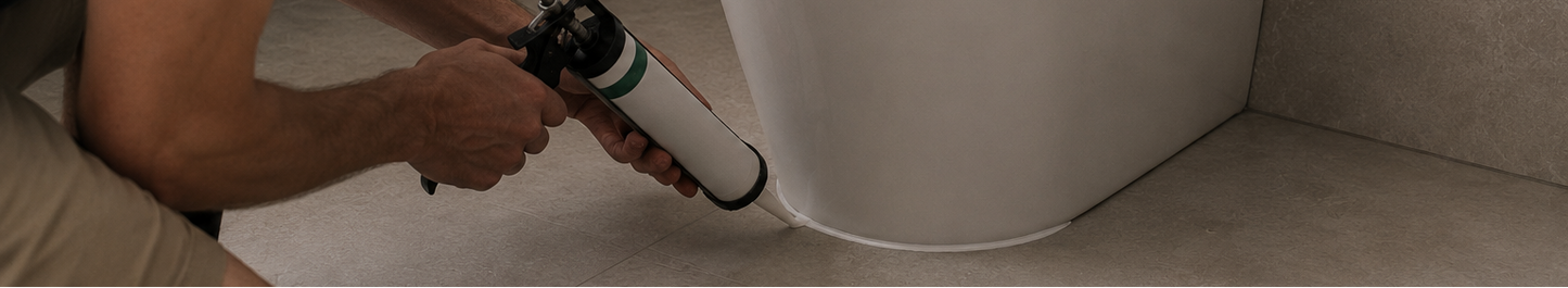 A man kneels beside a white toilet in a bathroom, applying silicone sealant with a caulking gun. A roll of tape and a towel are on the floor nearby.